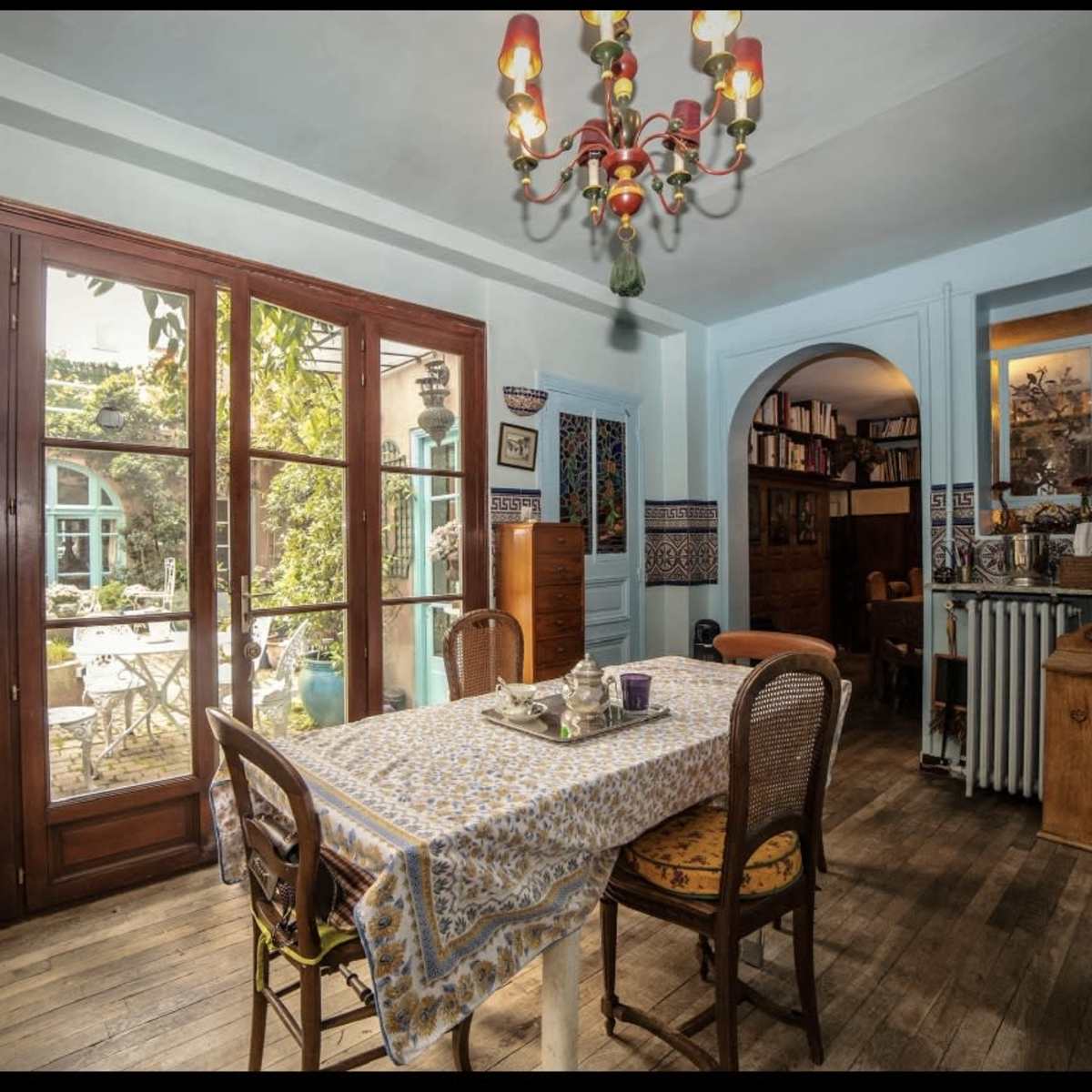 A spacious kitchen features a dining table set with a tablecloth, surrounded by chairs, with large windows showcasing a garden outside.