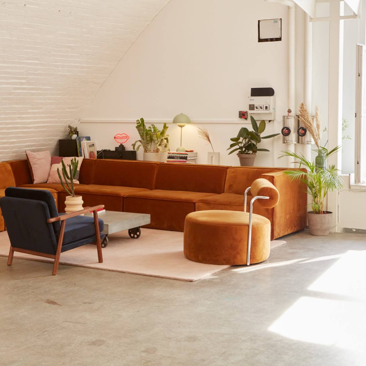 A modern living space featuring a large brown sectional sofa, a small round ottoman, a blue accent chair, and a coffee table on a light rug, all arranged under an arched ceiling with a large window.