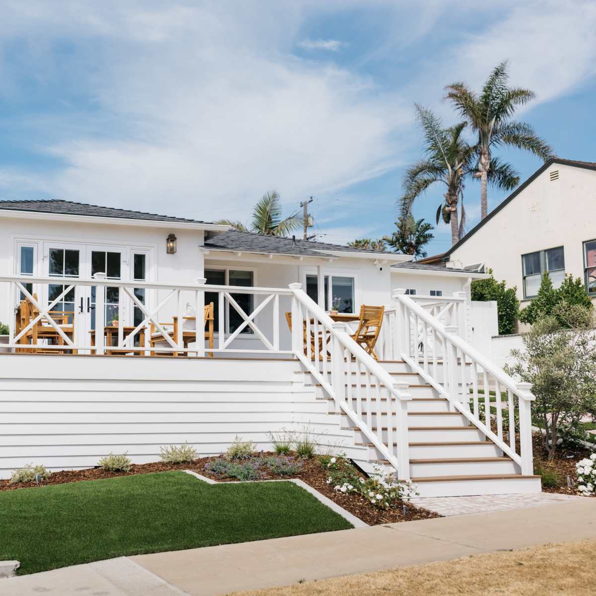 A single-story white house with a porch and staircase is situated on a corner lot, surrounded by landscaped greenery and palm trees.