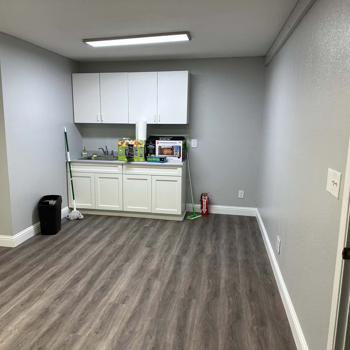A clean, empty room with a kitchenette featuring white cabinets and a countertop, along with a mop, a trash can, and a fire extinguisher.