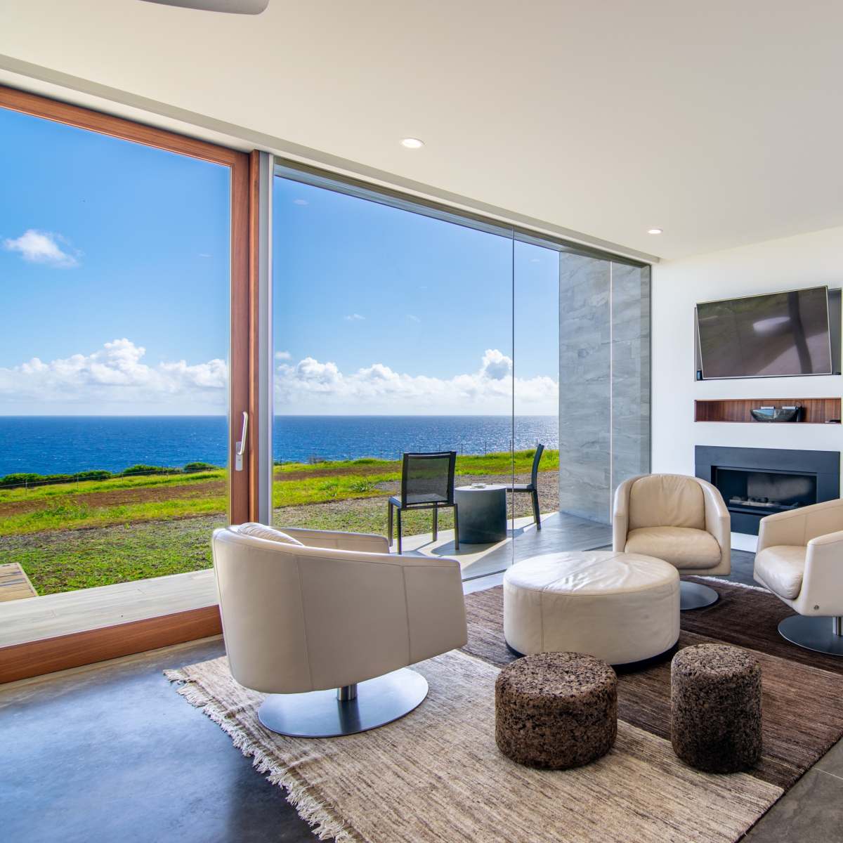 The scene depicts a modern living room with two white swivel chairs, a circular coffee table, a television mounted on the wall, and large windows offering a view of the ocean and sky.