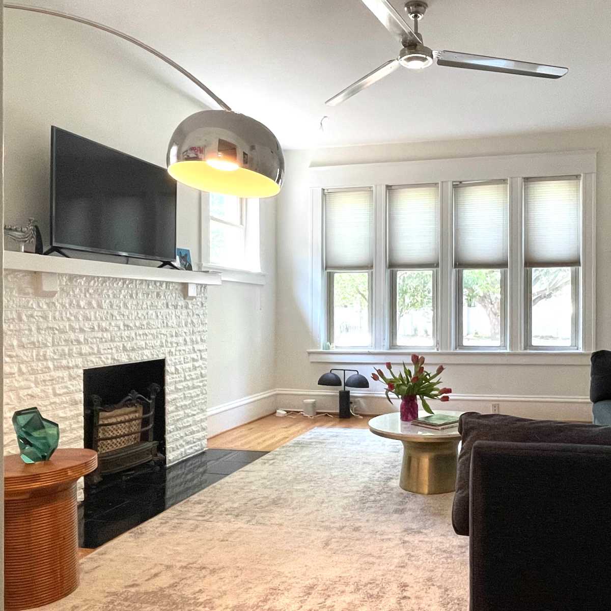 A cozy living room featuring a sectional sofa, a round coffee table with a floral arrangement, a modern lamp, and a flat-screen TV mounted above a white brick fireplace.