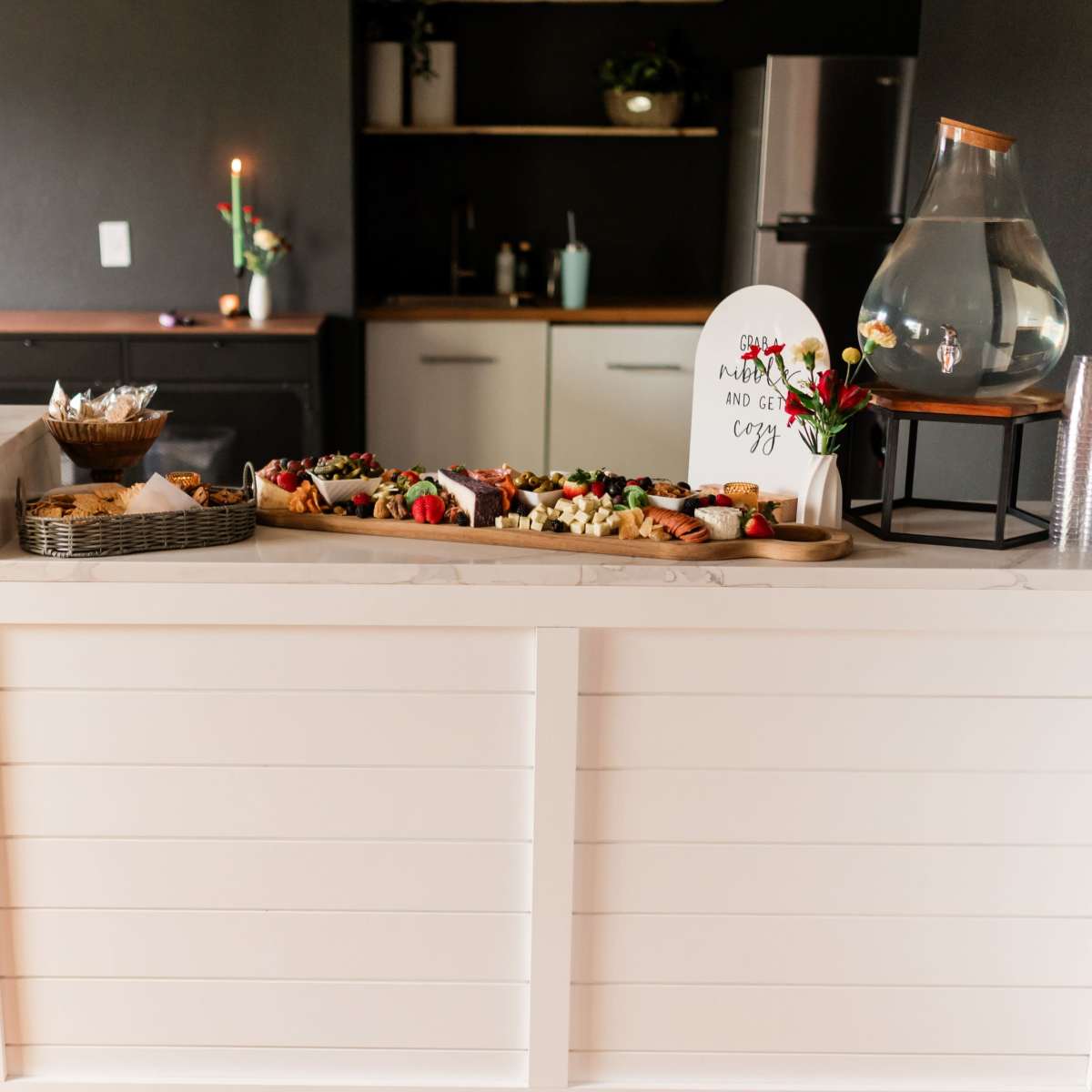 A refreshment station displays a variety of food items on a countertop, with a decorative sign and beverages in a clear container.