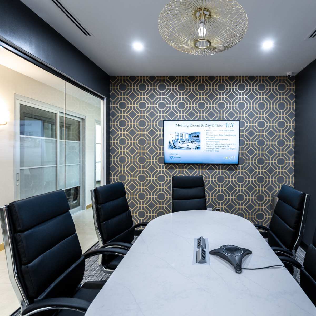 A modern conference room features a round table surrounded by black ergonomic chairs, with a display screen on the wall and patterned wallpaper in the background.