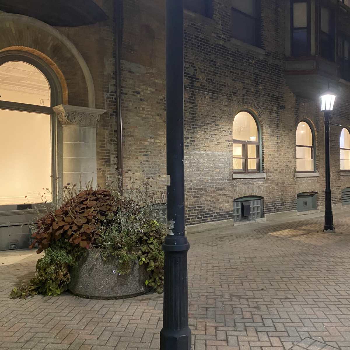 A brick pathway leads alongside a building with arched windows, illuminated by a streetlamp at night.