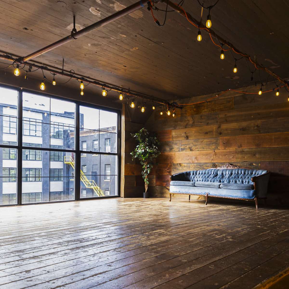 A large, open studio space with wooden floors, a blue sofa, and string lights hanging from the ceiling, illuminated by natural light from large windows.