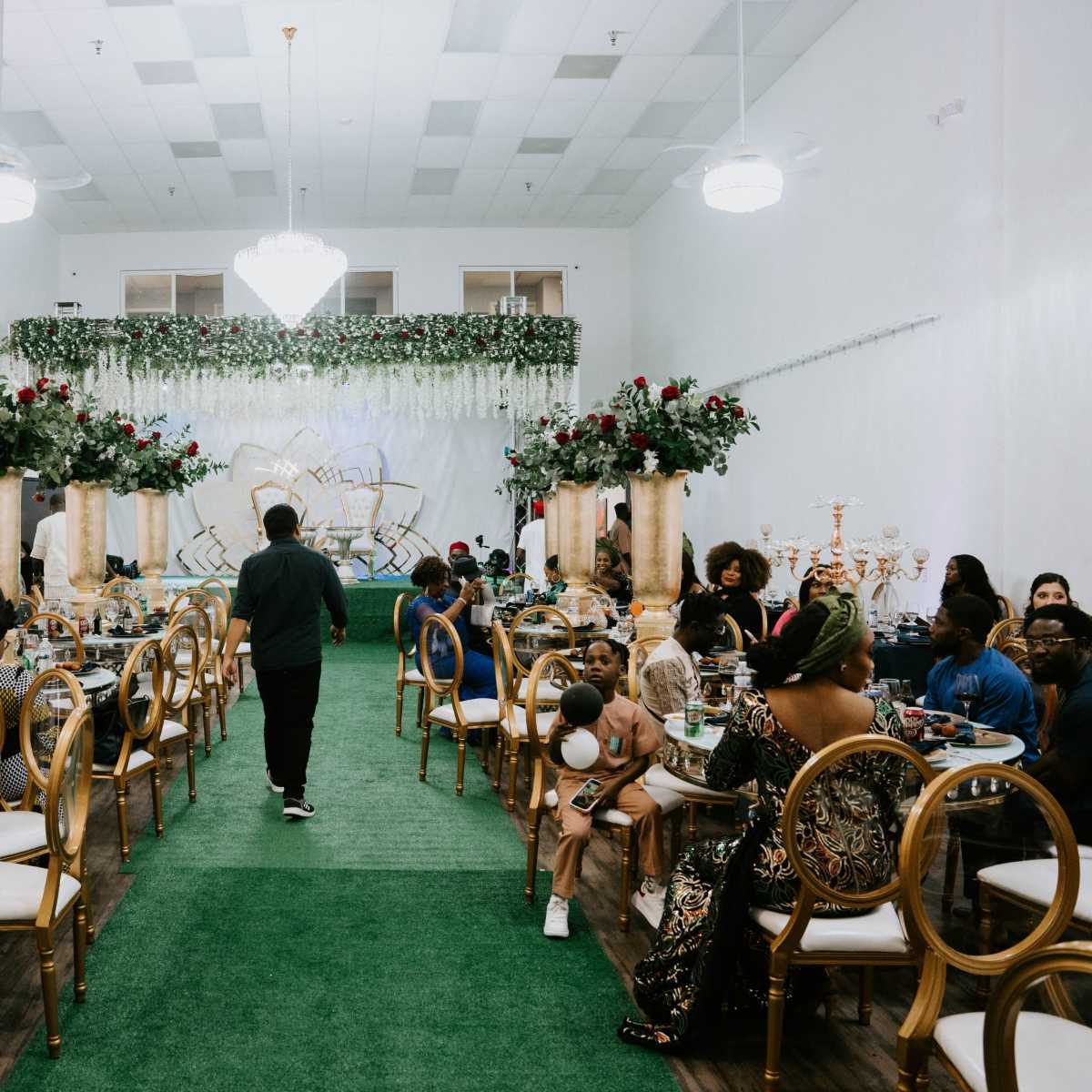 A banquet hall is set up for a formal event, featuring tables adorned with floral centerpieces, gold accents, and green carpet.