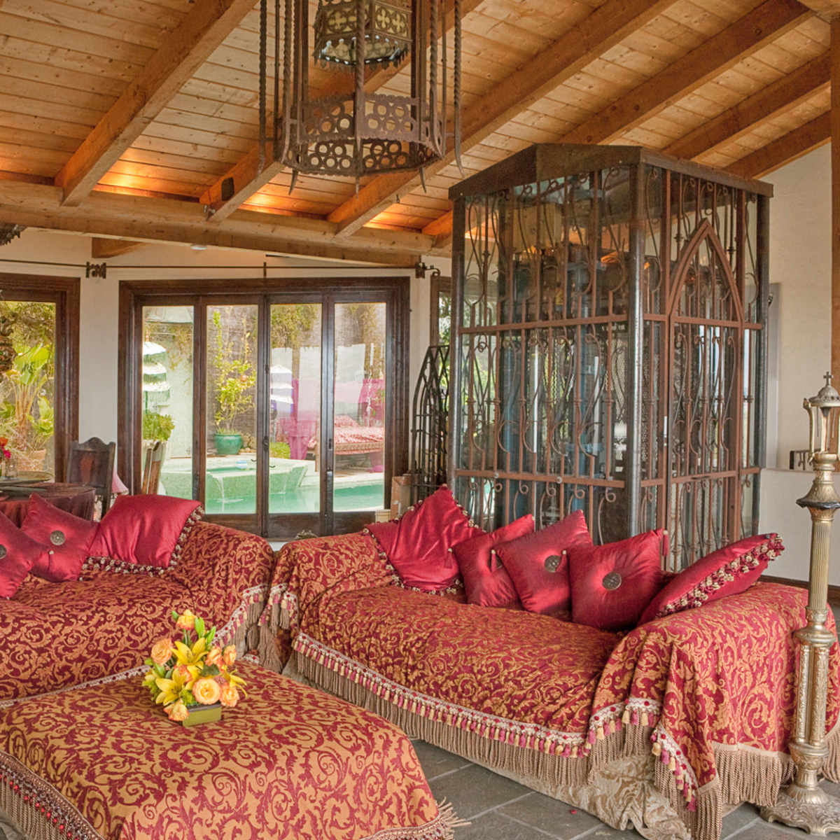 A spacious living room with wooden beams, large windows, and ornate red couches adorned with decorative pillows, set around a coffee table.