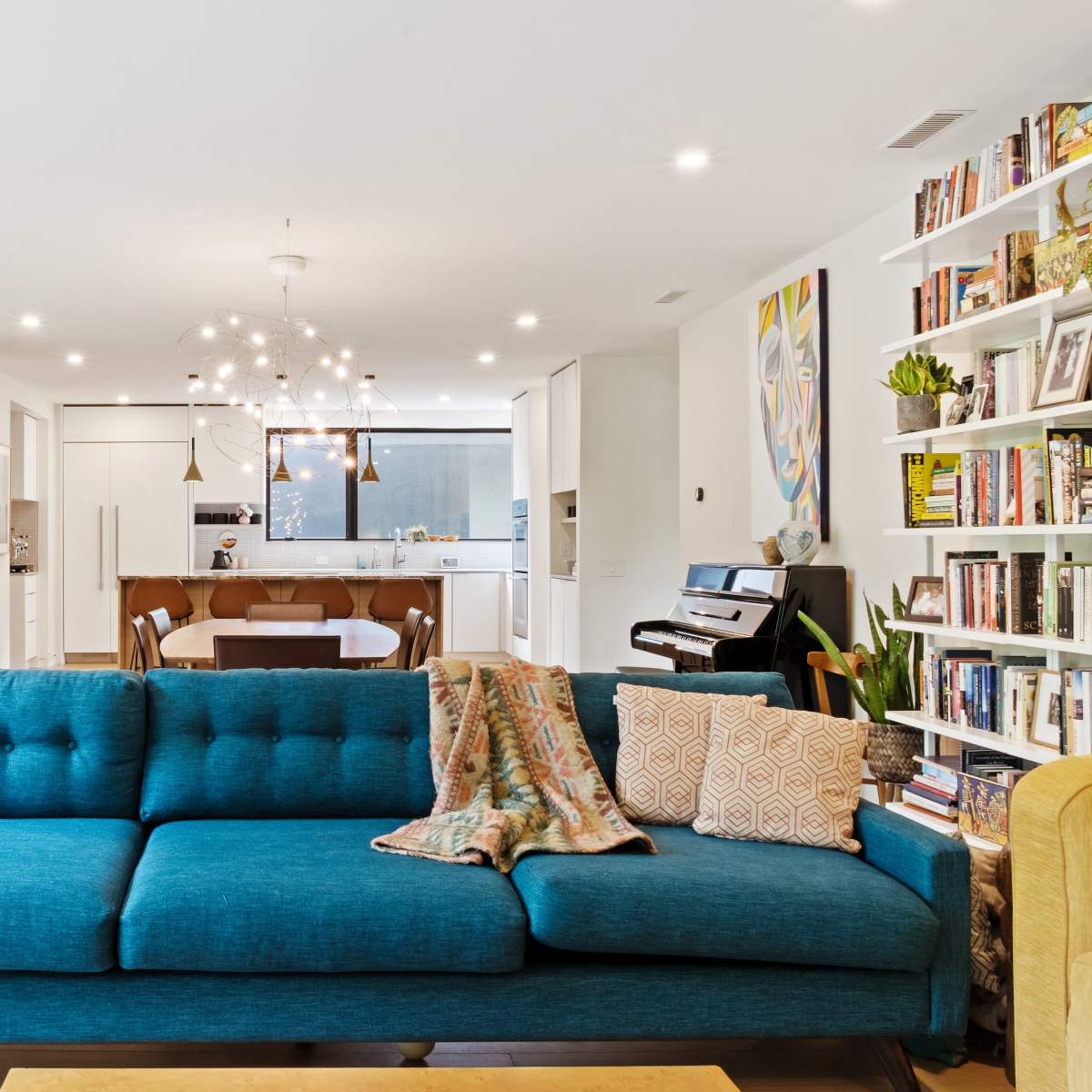 A bright living room features a blue couch with a patterned throw, alongside a bookshelf and an open kitchen area in the background.