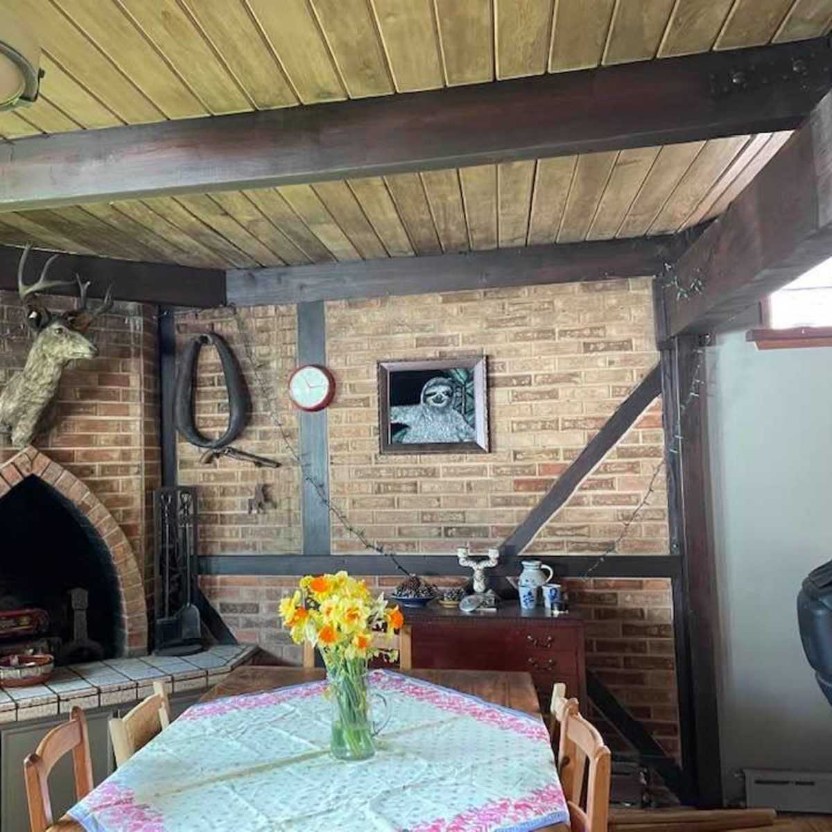 The interior features a wooden ceiling, a brick wall with a fireplace, a dining table adorned with flowers, and various wall decorations.