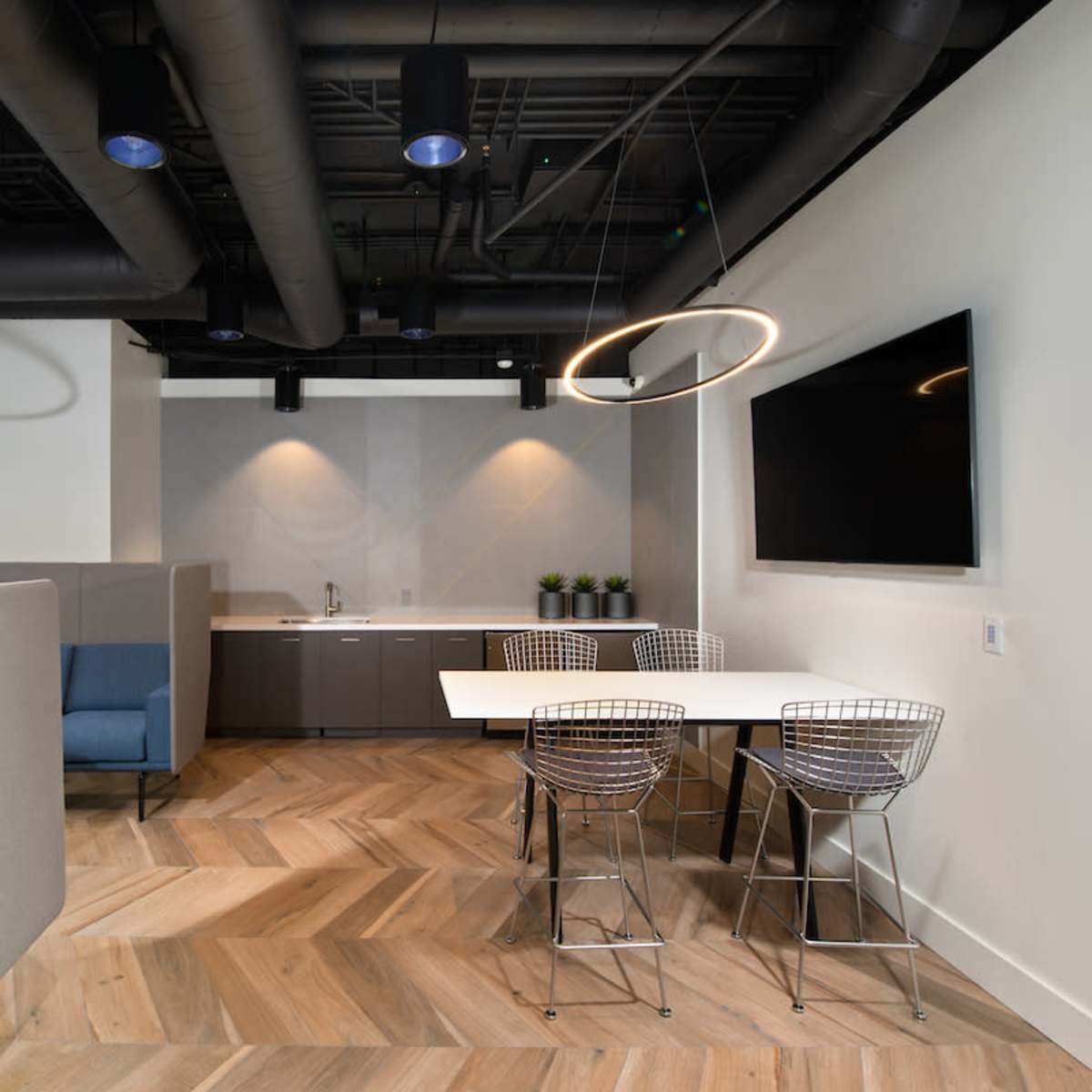A modern office space featuring a kitchenette, a table with wire chairs, and soft seating areas under circular pendant lights.