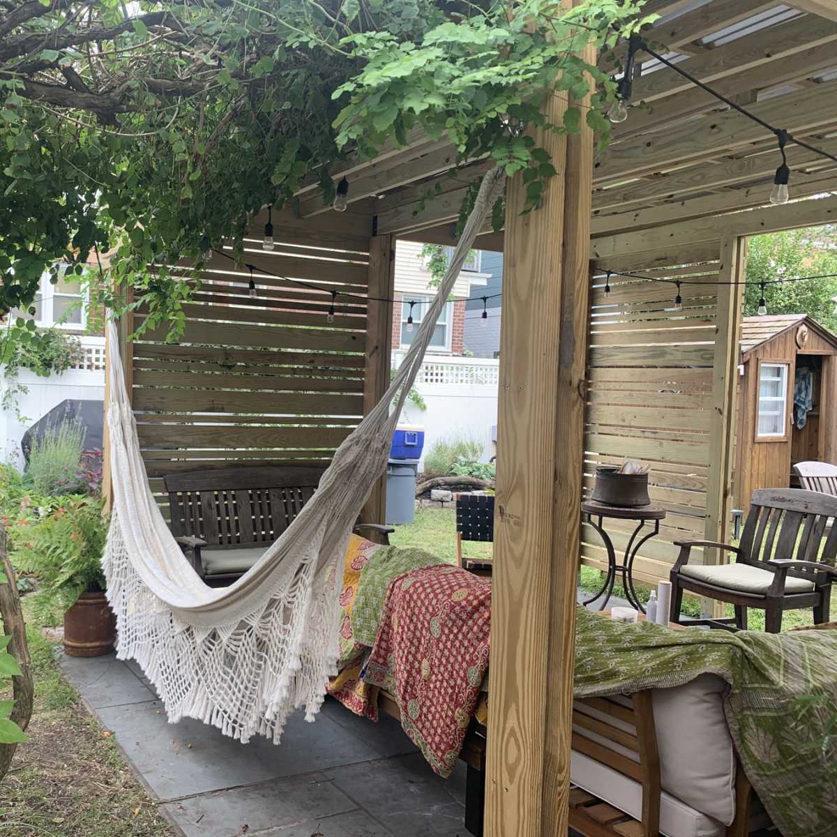 A hammock is suspended between wooden posts under a leafy pergola in a garden setting.