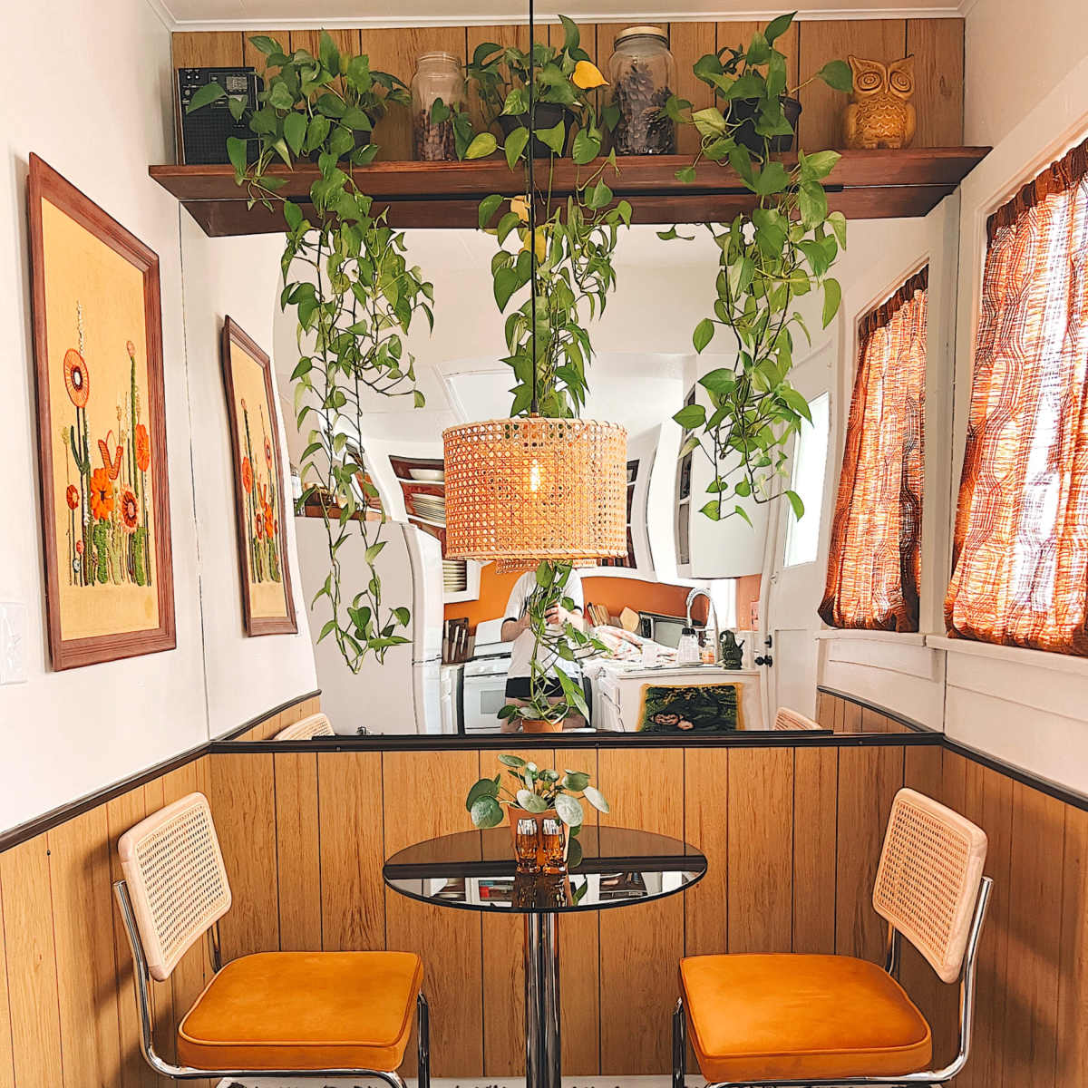 A small dining area features two orange chairs at a glass table, with hanging plants and framed floral artwork on the walls.