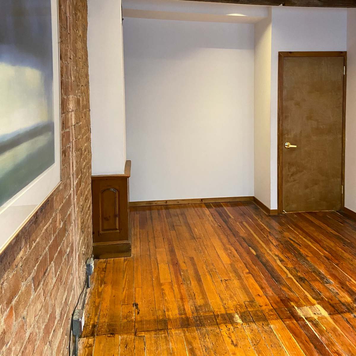 A room with exposed wooden beams, a brick wall, and polished hardwood flooring, featuring a blank wall and a closed door.
