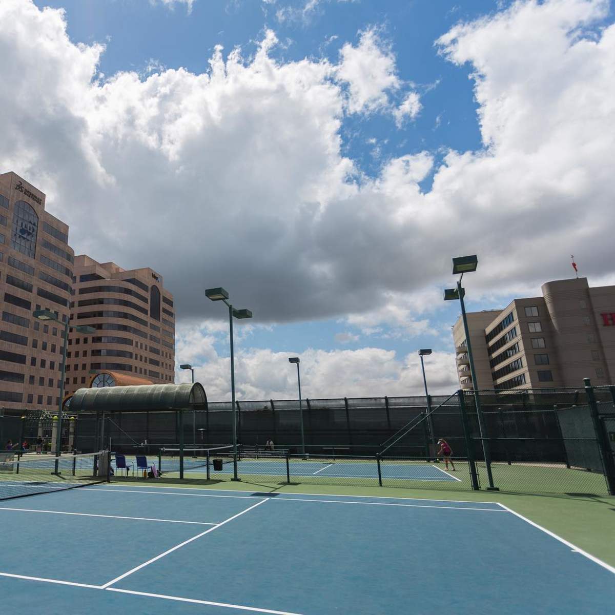 A tennis court surrounded by buildings and partly cloudy skies.