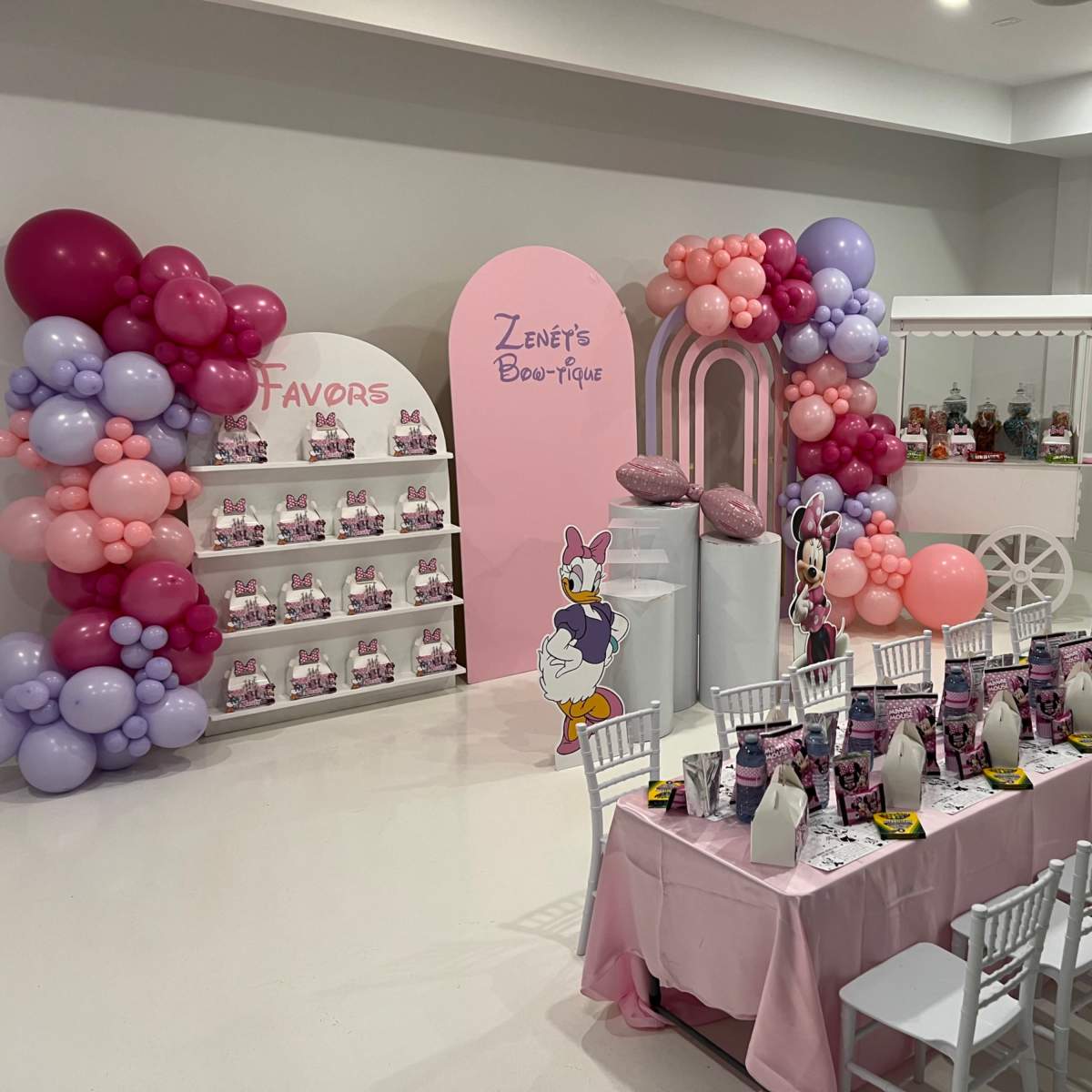 A decorated event space featuring a long table set for a celebration, with a backdrop of colorful balloons and a display of favor bags.