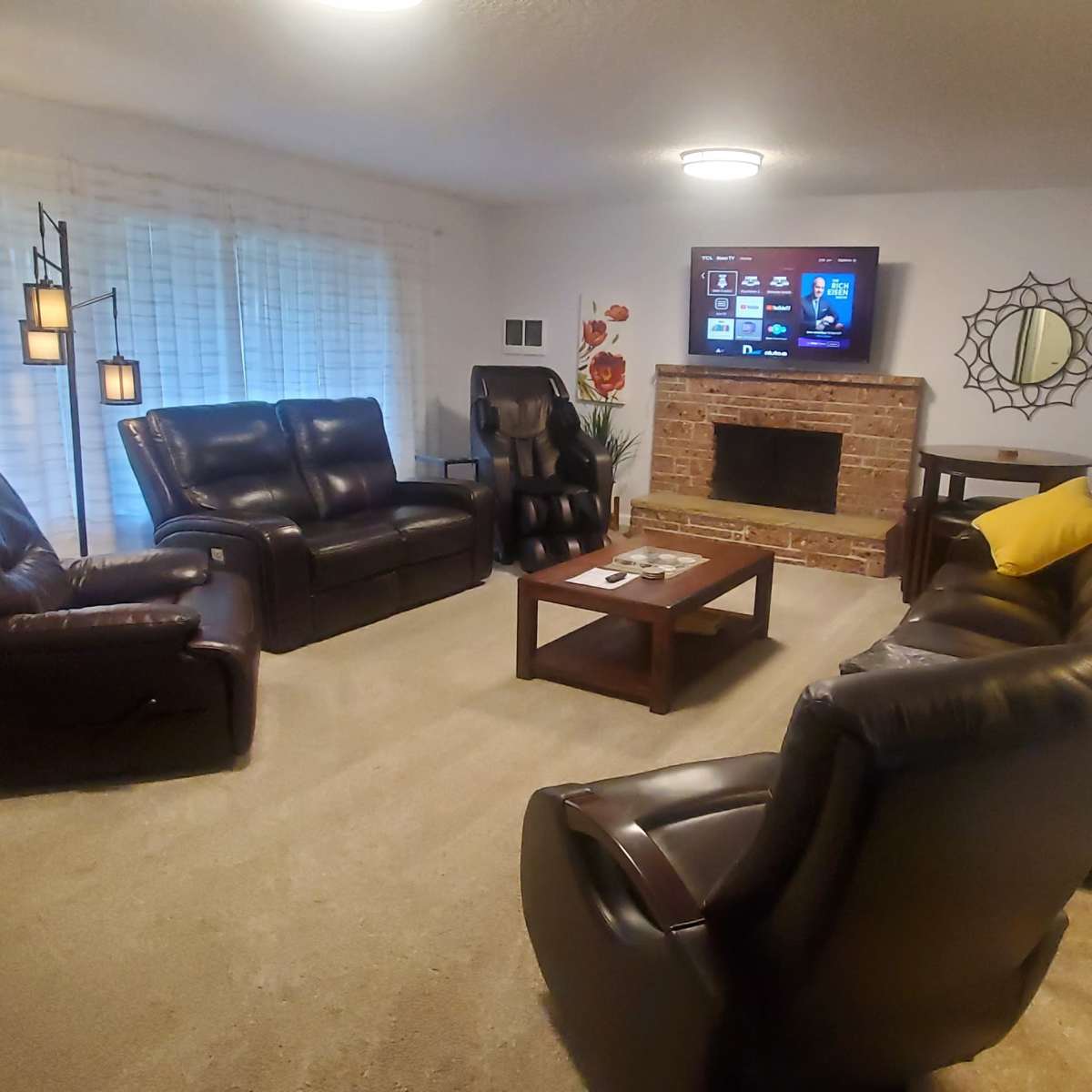 A living room features two recliners, a sofa, a coffee table, and a television mounted above a brick fireplace, with a large window covered by light curtains.