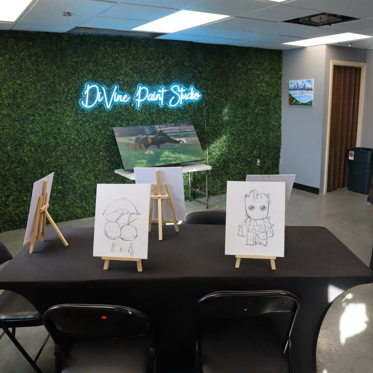 A room labeled "Divine Paint Studio," featuring a green wall with neon signage, a television, and several easels with drawings on display arranged around black tables.