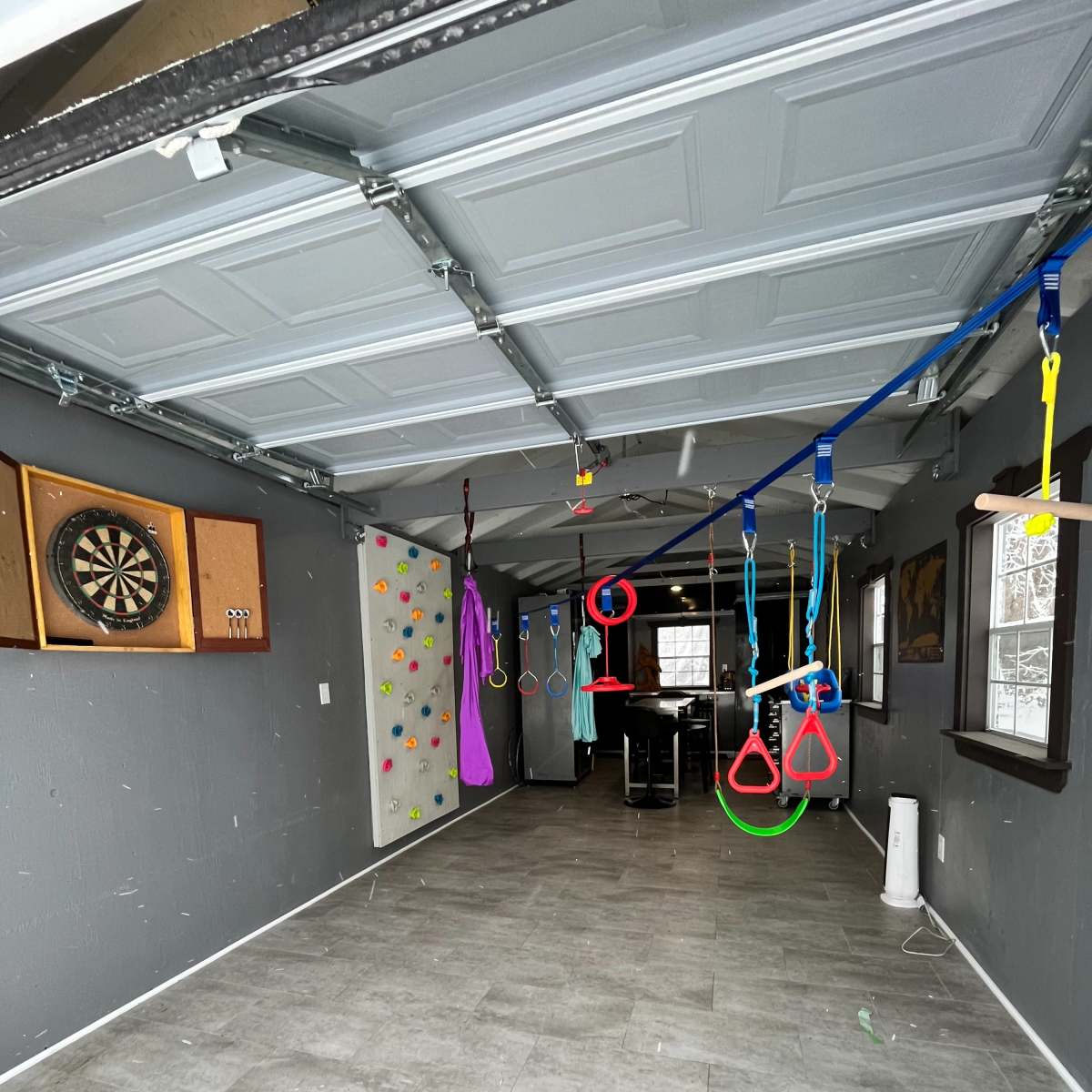 A garage space with a climbing wall, a dartboard mounted on the wall, and various gymnastic rings hanging from the ceiling.