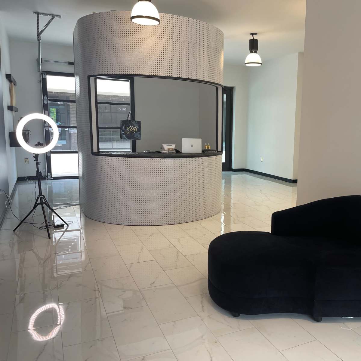 A modern interior space featuring a curved reception desk, a black chaise lounge, and a ring light setup on a polished tiled floor.