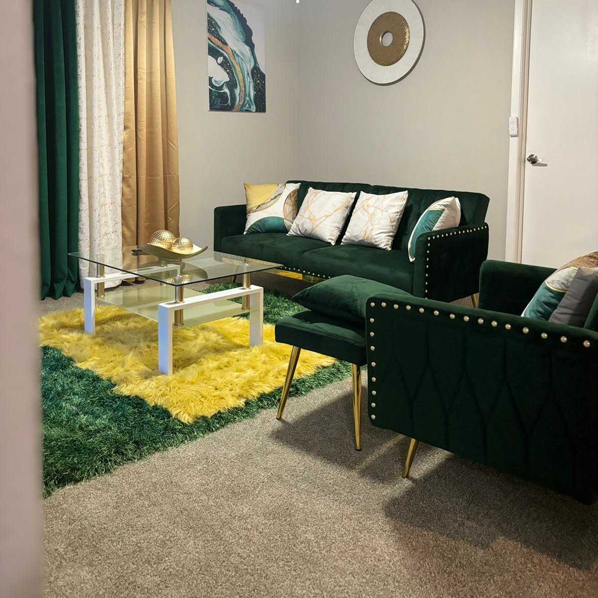 A modern living room furnished with a green sofa, a glass coffee table, and a yellow shag rug, with decorative curtains and wall art in the background.