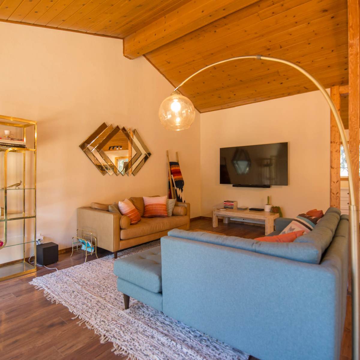 The living room features a beige sofa, a light blue sectional, a gold shelving unit, and a large wall-mounted television, all under a wooden ceiling.
