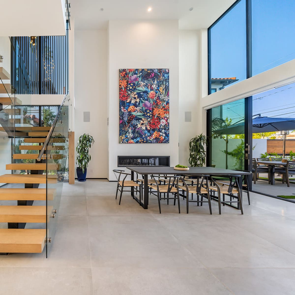 A modern interior featuring a staircase with wooden steps and a glass railing, alongside a dining area with a large table and a colorful wall artwork.