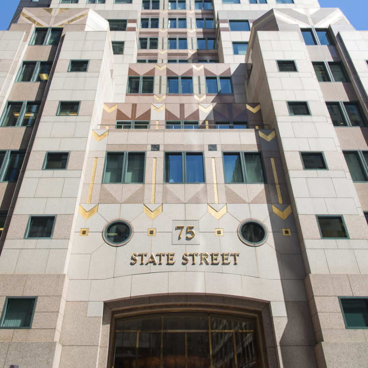 A tall modern building with a distinct architectural design located at 75 State Street, featuring a mix of glass and stone facades under a clear blue sky.