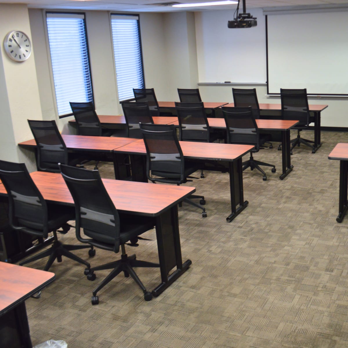 A classroom with several rows of desks and chairs arranged for seating, featuring a whiteboard and projector screen at the front.