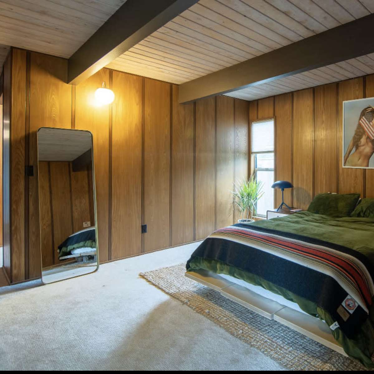 A bedroom with wooden paneling, a green bedspread, a mirror, and a lamp, creating a cozy atmosphere.
