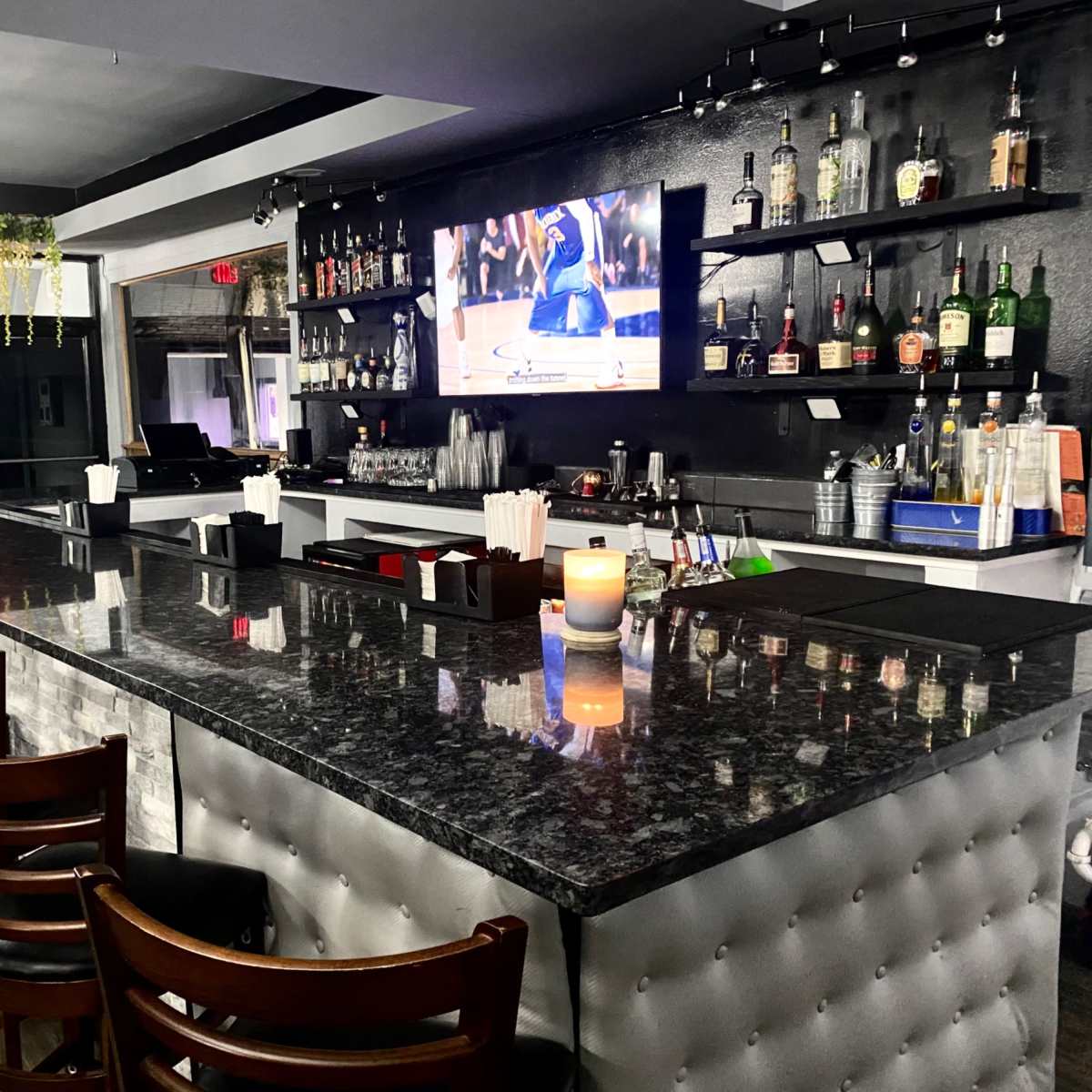 A visually organized bar area with a marble countertop, several bottles of liquor displayed behind, and a television screening a sports event.