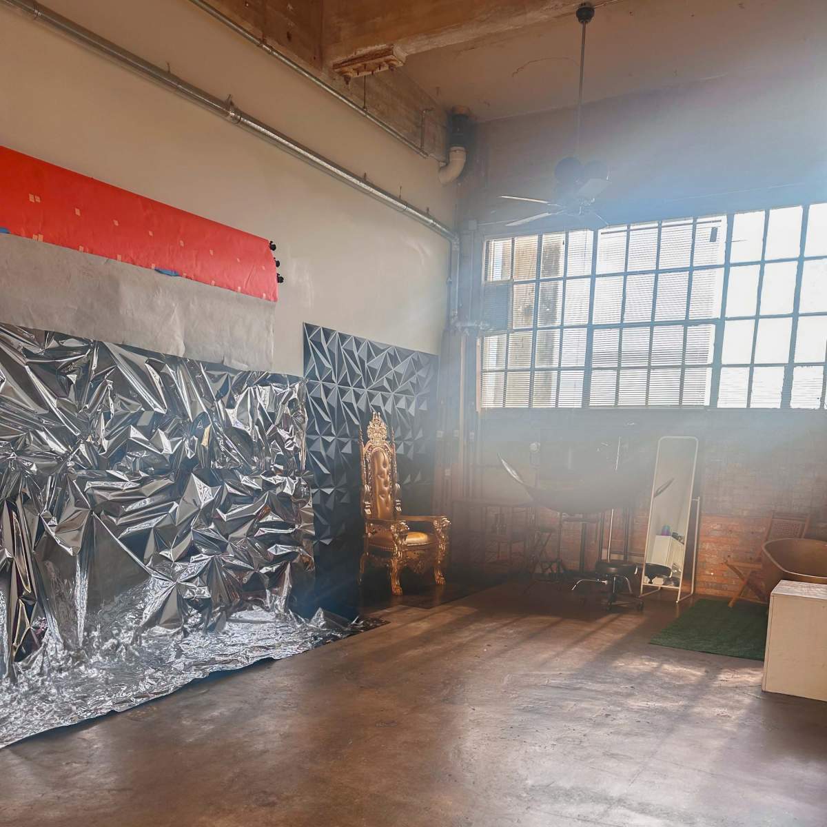 An industrial-style room with a large window, a golden chair, and a wall covered in reflective silver material.