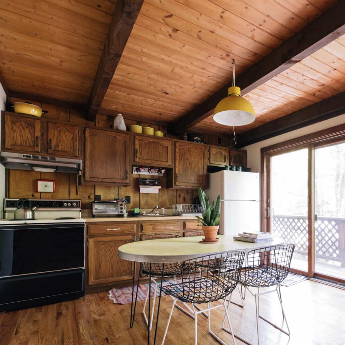 A wooden kitchen features brown cabinetry, a round dining table with wire chairs, and large windows overlooking a deck.