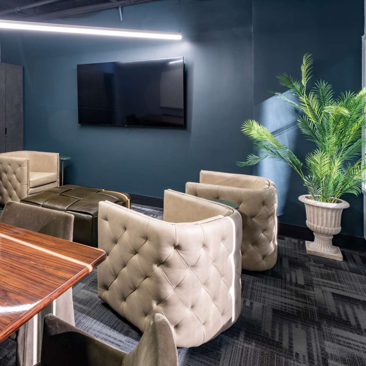 A modern meeting room with a polished wooden table, eight tufted chairs, a large television mounted on the wall, and a decorative plant in a corner.