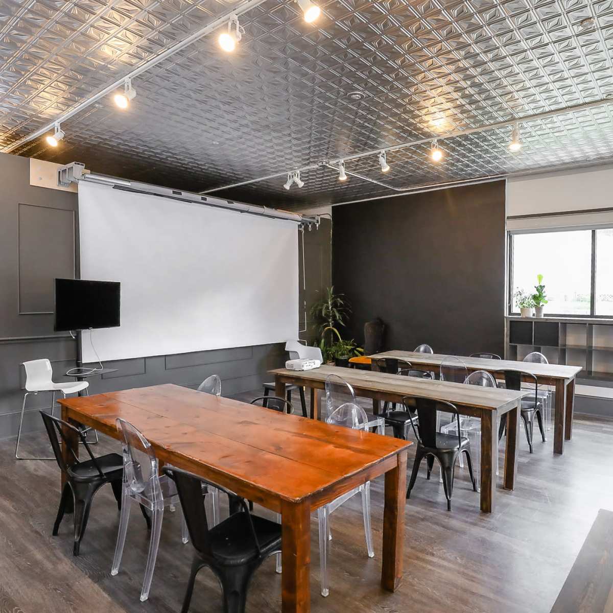 A modern meeting room with two long wooden tables, transparent chairs, a projector screen, and a TV on the wall.