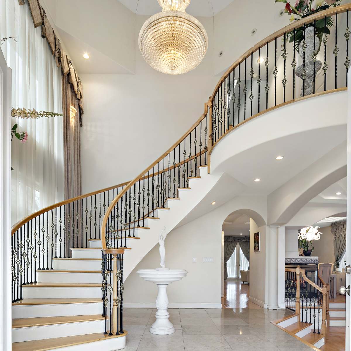 A grand entrance hall features a curved staircase with a wooden railing, a chandelier overhead, and a decorative fountain at the base.