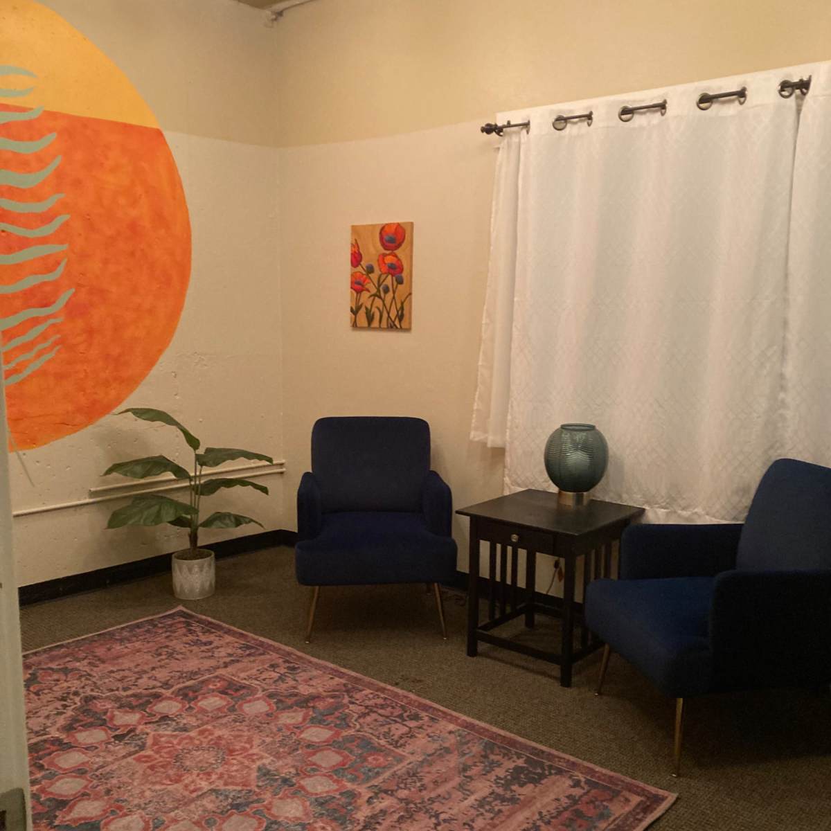 A small room with two blue chairs, a patterned rug, a table, a plant, and a large wall painting of an orange sun.