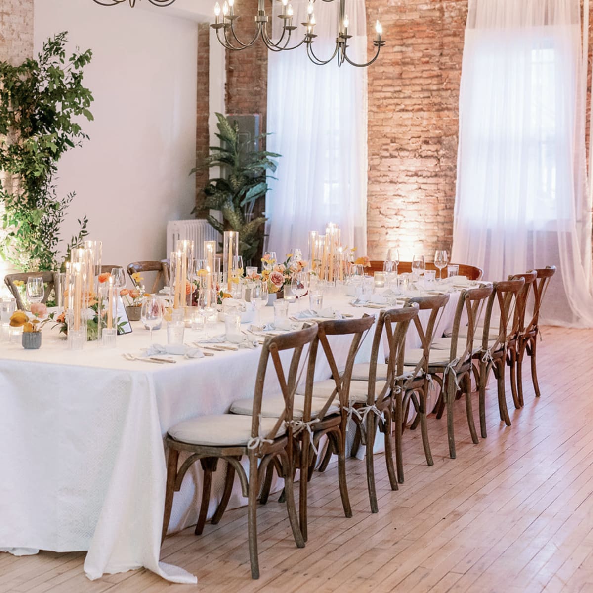 A long dining table is set with elegant tableware and surrounded by wooden chairs in a softly lit room with exposed brick walls.