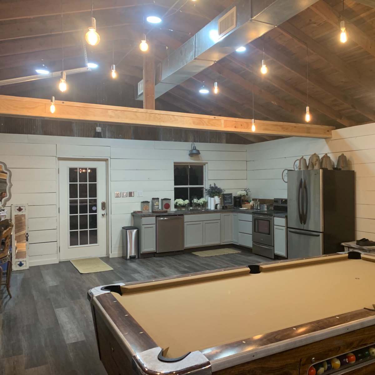A spacious room with a pool table in the foreground, wooden beams on the ceiling, and a kitchen area with modern appliances in the background.