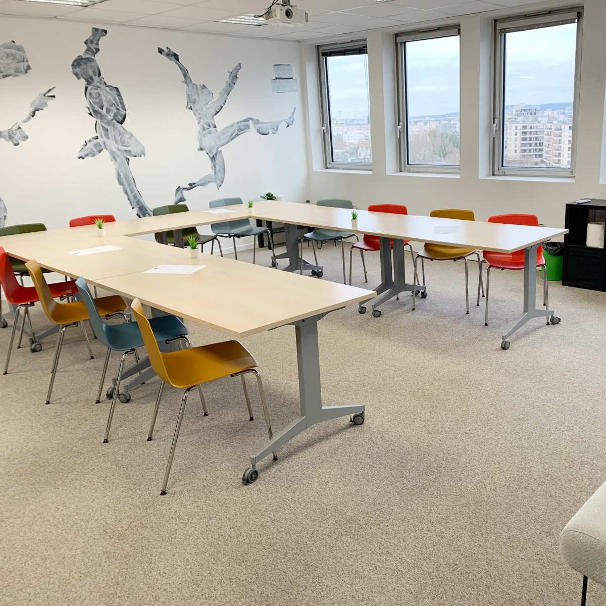A modern conference room with multiple tables arranged in a U-shape, colorful chairs, and large windows overlooking a cityscape.