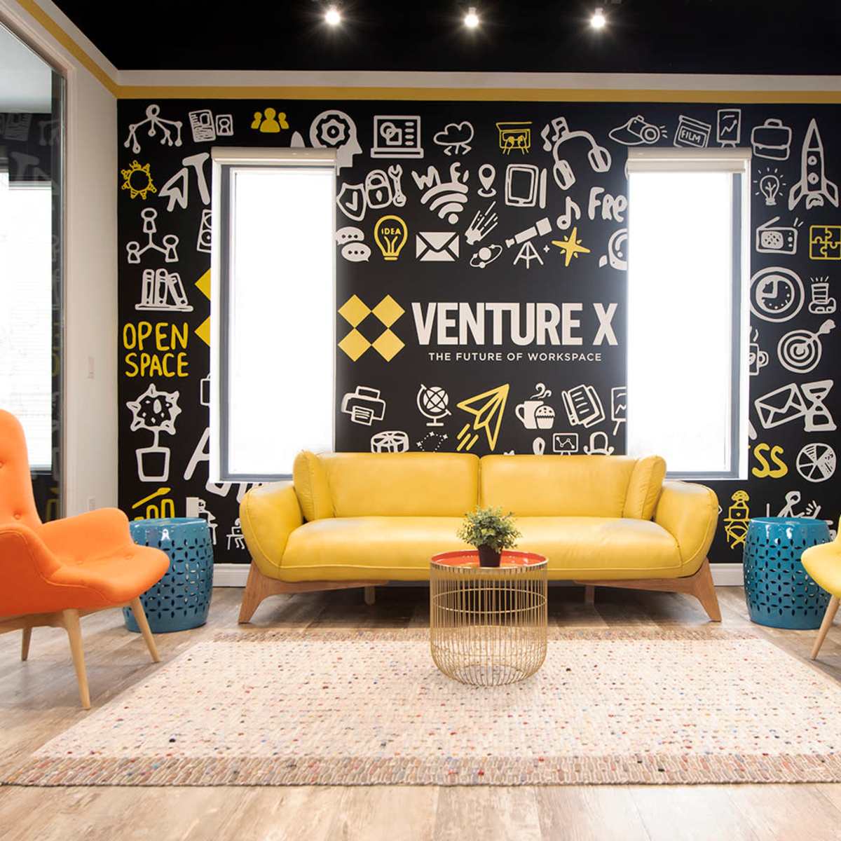 A modern workspace featuring a yellow couch, orange and yellow armchairs, a round coffee table, and a wall adorned with various black and white icons related to business and innovation.