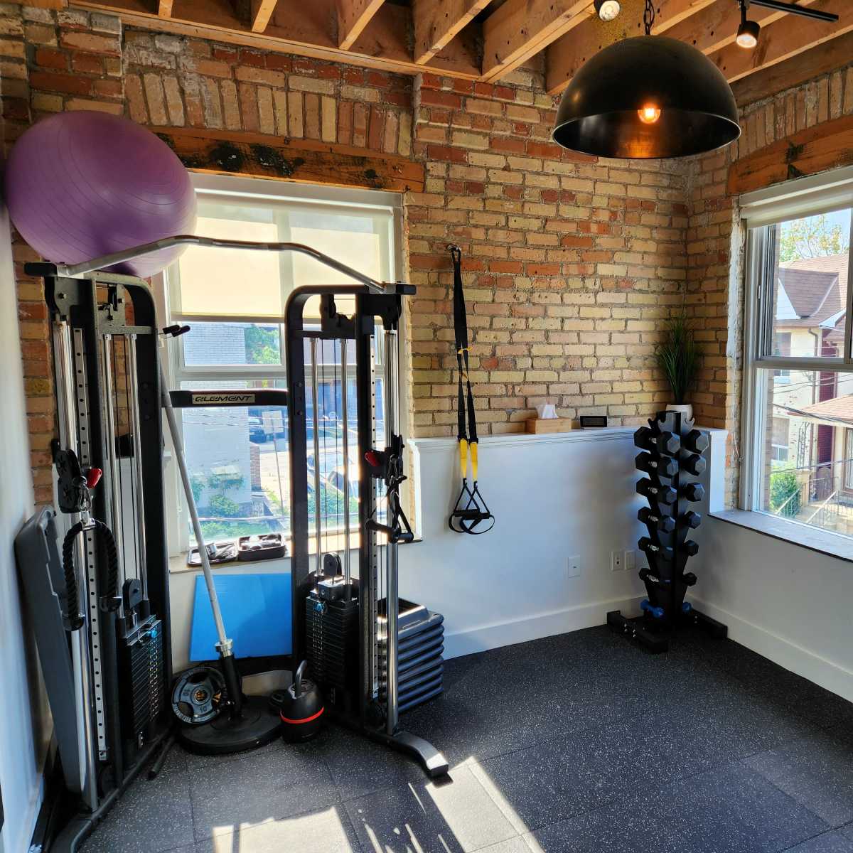 A home gym featuring exercise equipment, including a weight rack, a suspension trainer, and a stability ball, with large windows and exposed brick walls.