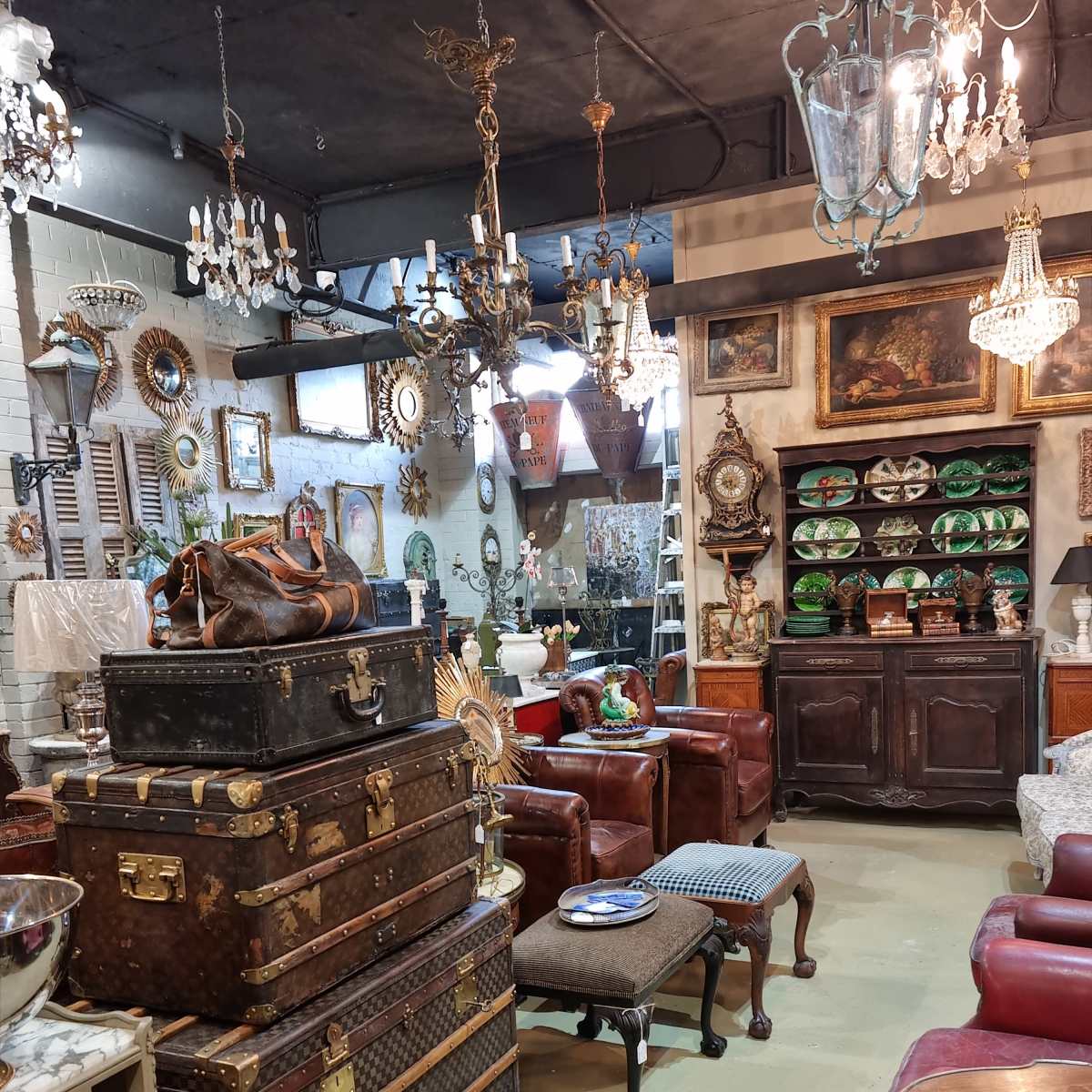 A vintage shop interior filled with various antique furniture, decorative items, and chandeliers, arranged around leather sofas and stacked trunks.