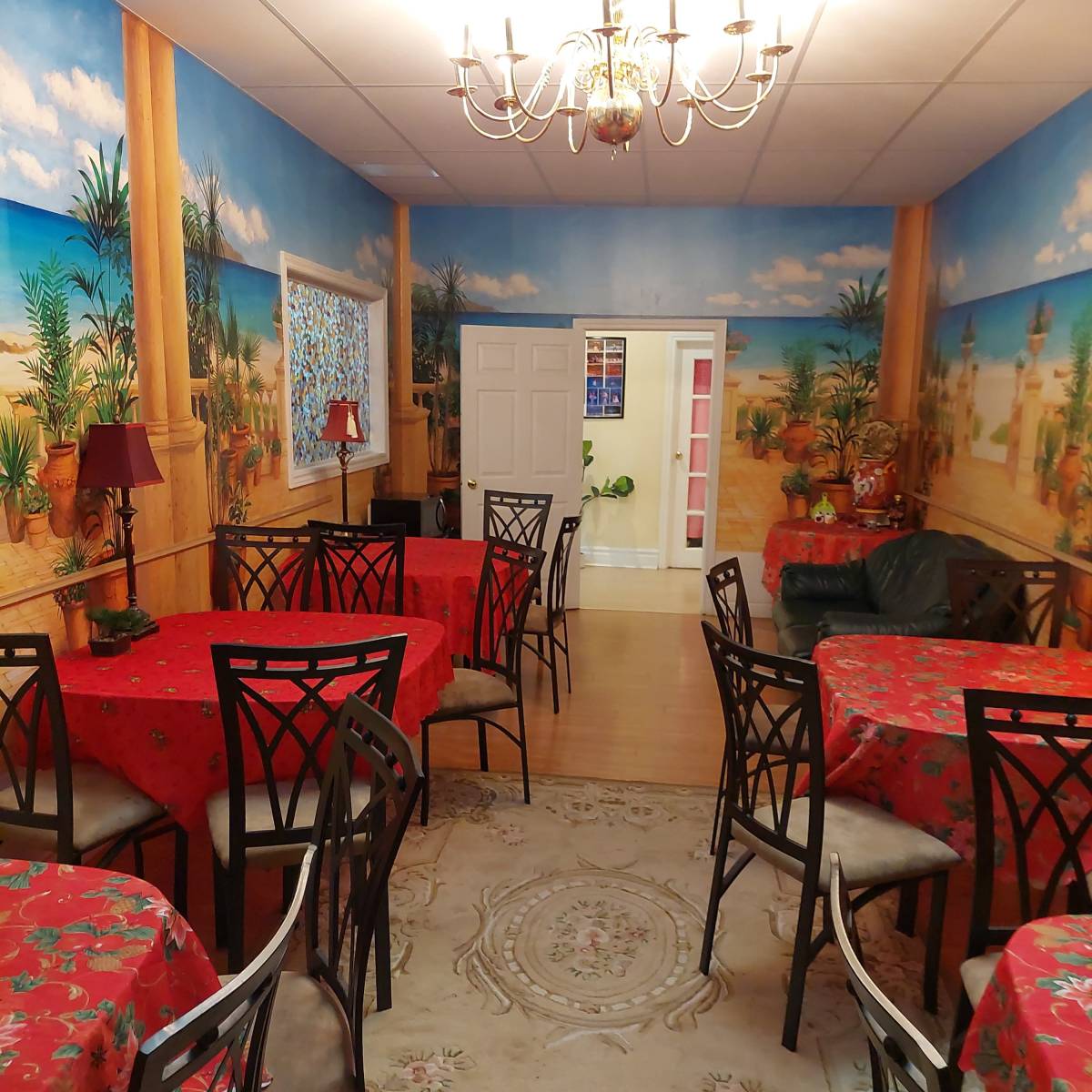 A dining area with several tables covered in red tablecloths, surrounded by black chairs, and walls decorated with tropical scenery murals.