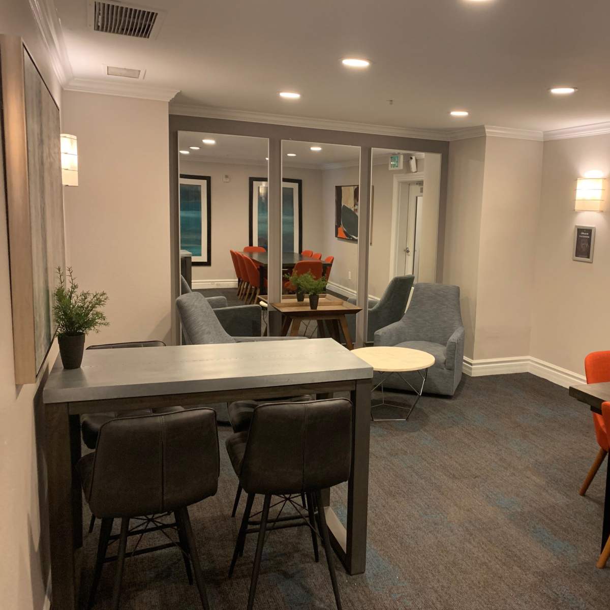 A modern lounge area featuring gray seating, a small round table, and a dining space with orange chairs.