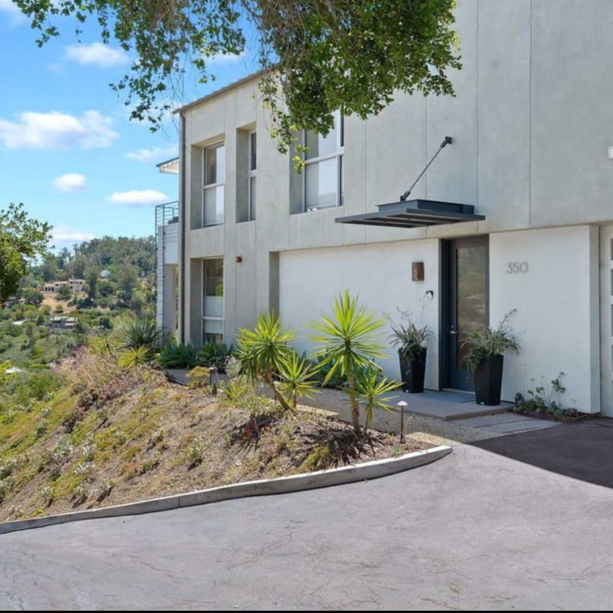 A modern two-story house with a concrete exterior and a sloped driveway is surrounded by greenery and offers a view of the landscape beyond.