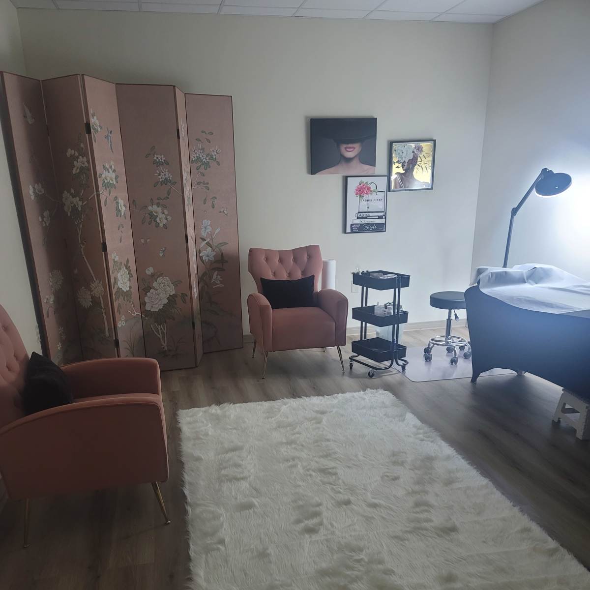 A softly lit room featuring two pink chairs, a decorative folding screen, a treatment table with a stool, and a large white rug.