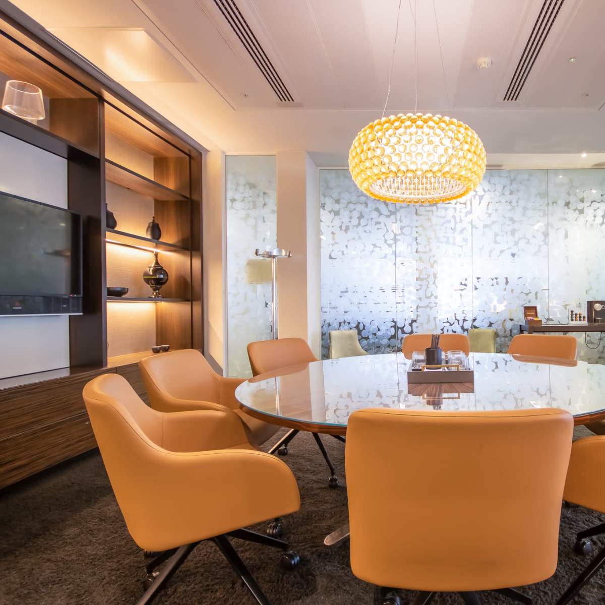 A modern conference room features a round glass table surrounded by orange chairs, with a decorative pendant light and frosted glass walls.