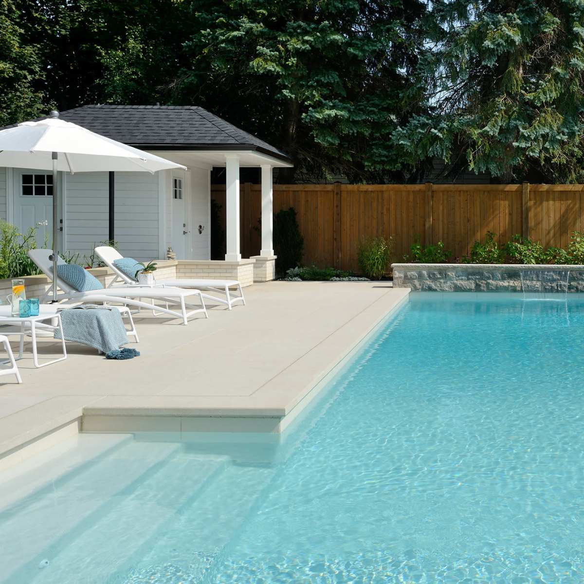 A clear swimming pool surrounded by lounge chairs and an umbrella, with a gazebo and wooden fence in the background.