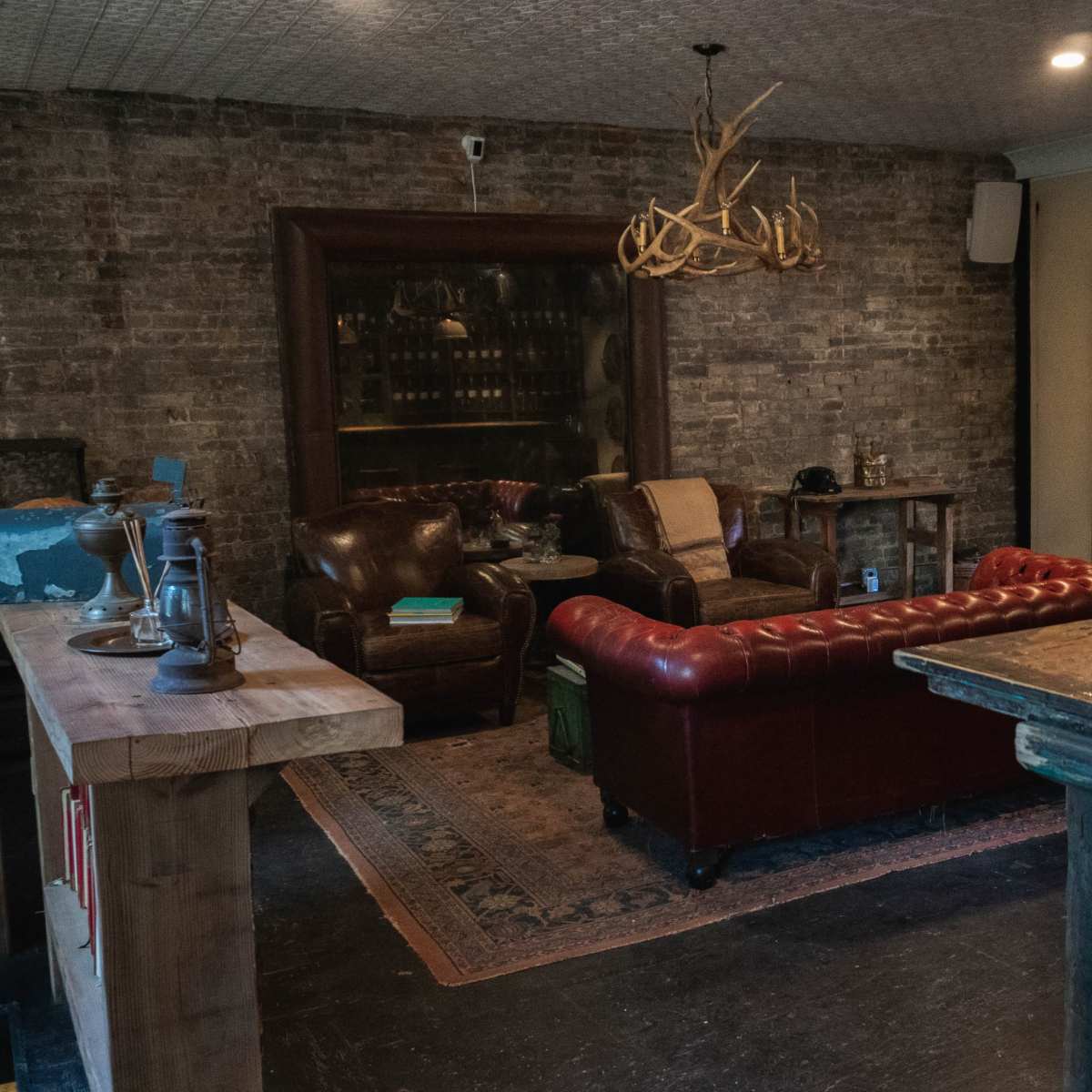 A cozy lounge area with leather armchairs, a tufted red sofa, a wooden table, and exposed brick walls.